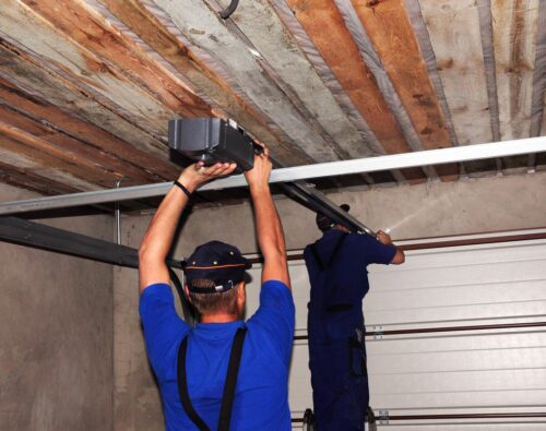 Two workers in blue uniforms install or repair a garage door opener mechanism on the ceiling of a garage with exposed wooden beams.