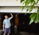 A man in casual clothes lifts a partially open garage door, with a car and various items visible inside the garage; green leaves are in the foreground.