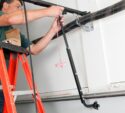 Person on a red ladder repairs or installs a garage door spring mechanism, using tools on the garage ceiling.