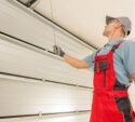 A technician in red overalls inspects or repairs a raised garage door, holding a cable and looking up at the mechanism.