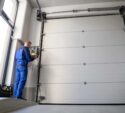 A technician in blue overalls operates a control panel next to a large closed industrial garage door inside a building.