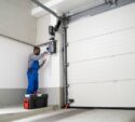 A technician in blue overalls repairs or inspects a garage door opener, standing on a small platform next to a toolbox in a modern garage.