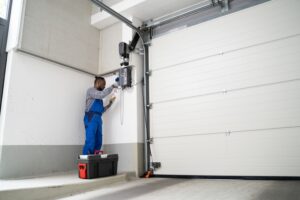 A technician in blue overalls repairs or inspects a garage door opener, standing on a small platform next to a toolbox in a modern garage.