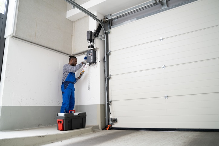 A technician in blue overalls repairs or inspects a garage door opener, standing on a small platform next to a toolbox in a modern garage.