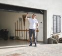 Man in casual clothes standing in front of a half-open garage door, looking up while holding the door; various tools and firewood are visible inside and beside the garage.
