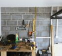 A workbench in a garage with various tools, glue, paint cans, and a spirit level on a brick wall.