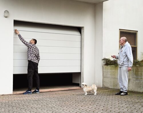 A man opens a garage door while another man stands nearby holding a remote control, and a small dog stands on the driveway.