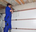 A person in blue overalls stands on a ladder, using a power drill to install or repair a garage door mechanism.