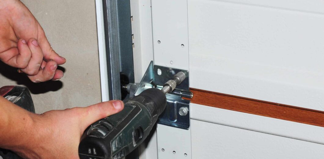 Person using a power drill to fasten or unfasten a screw on a metal bracket attached to a garage door.