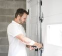 A man in a white shirt uses a tool to adjust the hardware on a white garage door inside a garage with cinderblock walls.