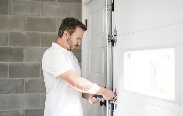 A man in a white shirt uses a tool to adjust the hardware on a white garage door inside a garage with cinderblock walls.