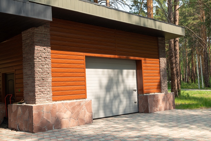A closed garage door on a modern building with wood and stone exterior, surrounded by trees and a paved driveway.