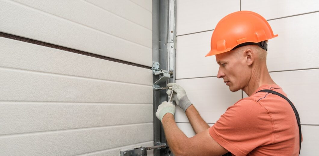 A worker in an orange hard hat and gloves uses a tool to fix or adjust a metal component on a garage door.