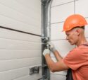 A worker in an orange hard hat and gloves uses a tool to fix or adjust a metal component on a garage door.