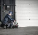 A person in work clothes and a cap kneels beside a closed garage door, working with a toolbox in an industrial setting.