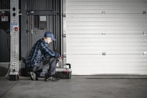 A person in work clothes and a cap kneels beside a closed garage door, working with a toolbox in an industrial setting.