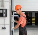 A worker in an orange hard hat and gloves is inspecting or repairing a garage door in a parking garage.