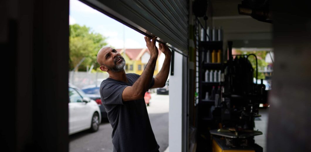 A man in a dark t-shirt pulls open a metal garage door at an automotive repair shop during the day. Cars and buildings are visible outside.