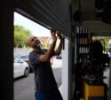A man in a dark t-shirt pulls open a metal garage door at an automotive repair shop during the day. Cars and buildings are visible outside.