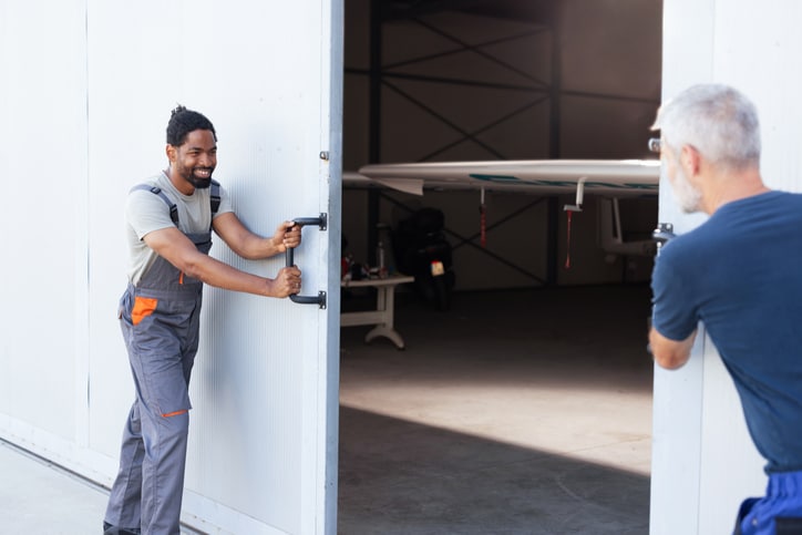 Two men open large hangar doors, revealing a small aircraft inside.