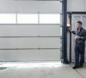 A man in work clothes operates a control on the wall to open or close a large industrial garage door inside a well-lit facility.