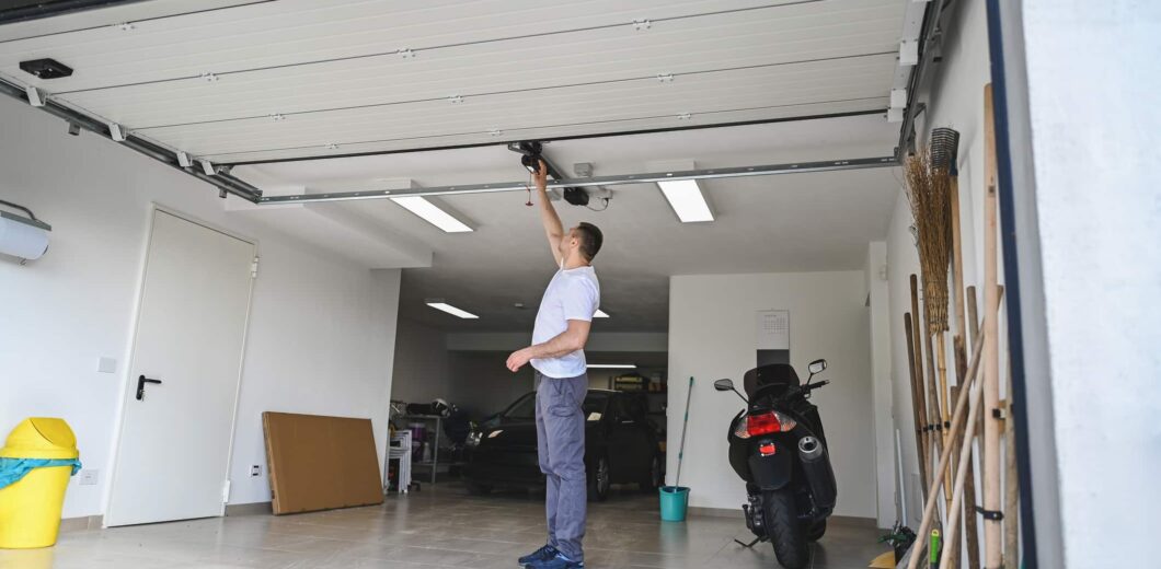 A person stands in a garage opening an overhead door by hand, with cars, a scooter, tools, and cleaning supplies visible inside.