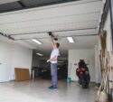 A person stands in a garage opening an overhead door by hand, with cars, a scooter, tools, and cleaning supplies visible inside.