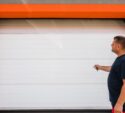A man stands in front of a closed white garage door with an orange frame, holding a remote control and facing the door.
