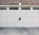 Closed beige garage door with three rectangular windows at the top, black handles in the center, and black decorative hardware, set in a brick and stone exterior wall.
