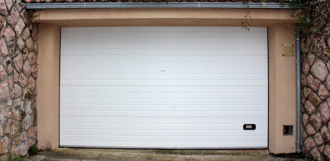 A closed white garage door set in a beige frame with stone walls on both sides; a small handle and a utility box are visible near the bottom right corner.