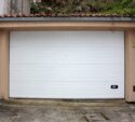 A closed white garage door set in a beige frame with stone walls on both sides; a small handle and a utility box are visible near the bottom right corner.