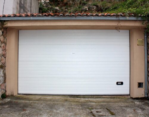 A closed white garage door set in a beige frame with stone walls on both sides; a small handle and a utility box are visible near the bottom right corner.
