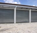 Three closed metallic roller garage doors set in a white building, with a concrete ground and a blue sky with clouds above.