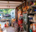 A cluttered garage with tools, bikes, shelves full of various items, and an open door showing vehicles and a trailer outside.