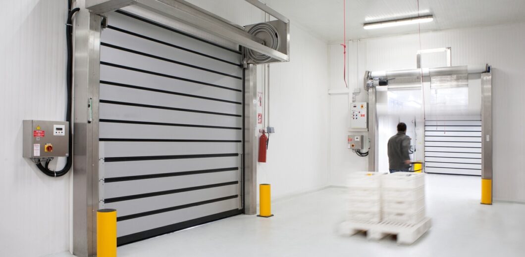 A person pushes a pallet jack with stacked trays through a clean, industrial loading area with large automated doors and white walls.