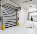 A person pushes a pallet jack with stacked trays through a clean, industrial loading area with large automated doors and white walls.