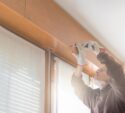 A person wearing gloves uses a measuring tape to measure a wooden panel above a window inside a bright room.