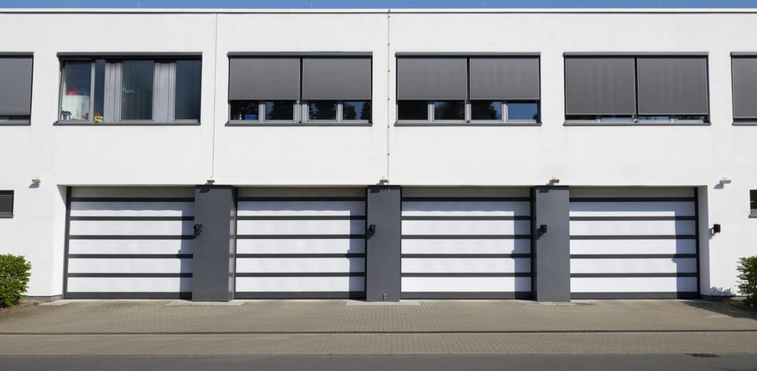 Four modern white garage doors with dark frames are aligned side by side in a white building, each with a window above, under a clear blue sky.