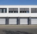 Four modern white garage doors with dark frames are aligned side by side in a white building, each with a window above, under a clear blue sky.