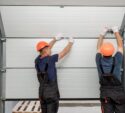 Two workers in safety helmets and overalls install or repair a sectional garage door from the inside of a building.
