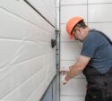 A man in work overalls and an orange hard hat repairs or installs a garage door, using a screwdriver on the metal frame next to a tiled wall.