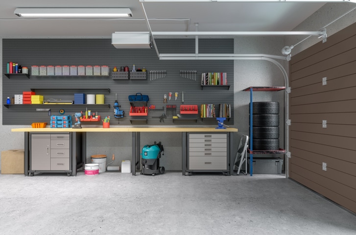 A clean, organized garage with workbenches, storage cabinets, tools on wall panels, a stack of tires, a vacuum, and the garage door partially open.