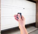 A hand holding a remote control in front of a partially open white garage door.