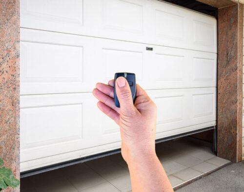 A hand holding a remote control in front of a partially open white garage door.