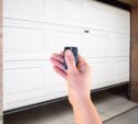 A hand holding a remote control in front of a partially opened white garage door with a stone and tile exterior.