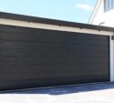 A closed, modern black garage door attached to a white building, with a paved driveway in front.