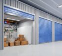 A row of storage units with blue doors; one unit is open, showing boxes, a mattress, and various tools and supplies organized on shelves.