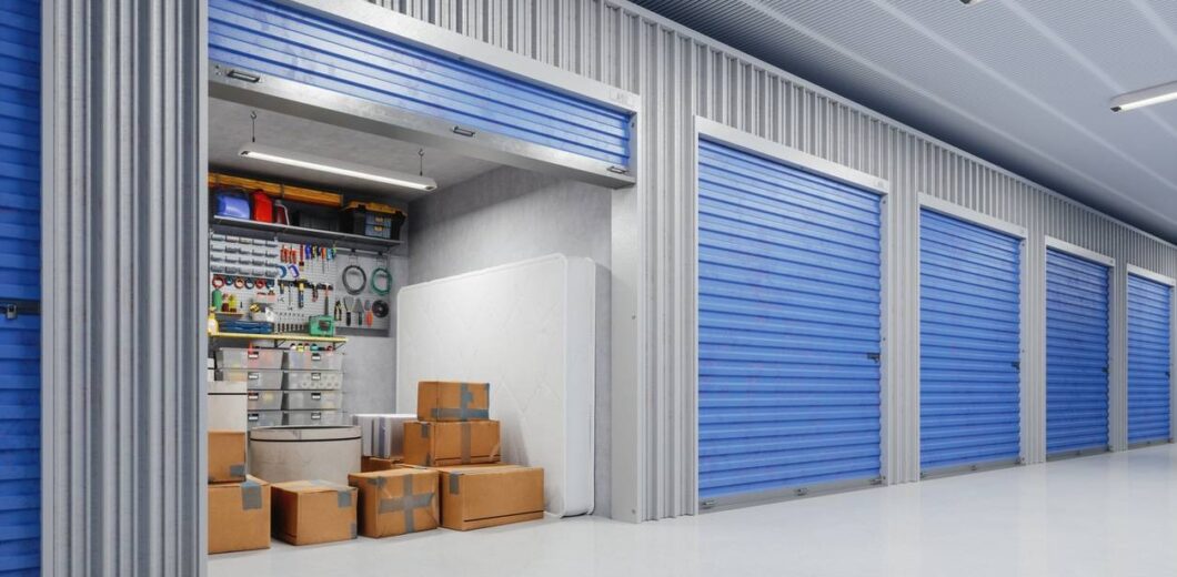 A row of storage units with blue doors; one unit is open, revealing cardboard boxes, a mattress, and organized tools hanging on the back wall.