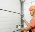 A worker in a hard hat and gloves is installing or repairing a sectional garage door, adjusting the metal tracks and hinges with a tool.