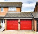Two red garage doors attached to a brick house with a tiled roof, a gravel driveway, and a small hanging flower basket between the doors.